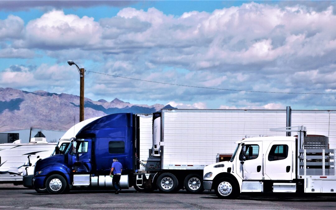 Two trucks, one white truck in front of a blue Semi-Truck with mountains in back.